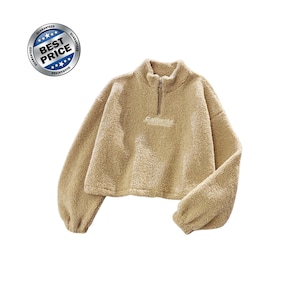 Könnte beinhalten: Ein beigefarbener, langärmeliger, bauchfreier Pullover aus flauschigem, strukturiertem Fleece-Material. Er verfügt über einen Stehkragen mit kurzem Frontreißverschluss und das Wort California, das in weißem Garn auf der Brust gestickt ist.