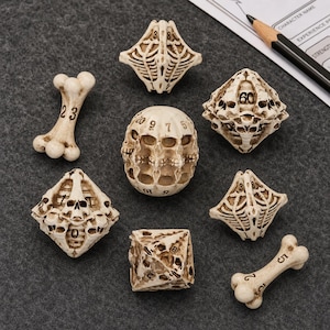 May include: A set of seven polyhedral dice and two bone-shaped dice with an off-white, aged bone finish. The dice feature intricate sculpted details including skulls and ribcage patterns, with black numerals clearly visible on each piece.