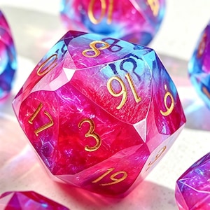 May include: A multi-sided, gemstone-like die with a vibrant blend of pink and blue hues. Gold numerals are visible on the facets. The die is designed for tabletop gaming.