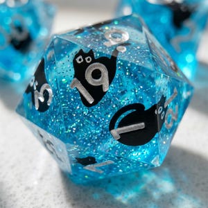 May include: A blue, glitter-filled, polyhedral die with black cat silhouettes and silver numbers. The die features the number 19 prominently. The dice are designed for tabletop games.