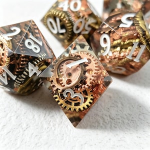 Puede incluir: Primer plano de varios dados poliédricos con temática steampunk. Los dados son transparentes con engranajes de cobre y latón incrustados en su interior. Los números blancos son visibles en las caras. Los dados están dispuestos sobre una superficie blanca.