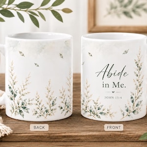 May include: A white ceramic mug featuring a botanical design of green and brown leaves at the base. The front displays the text Abide in Me. JOHN 15:4 in a dark script and serif font. The back shows a continuation of the leaf pattern.