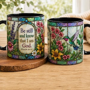 May include: Two ceramic mugs with black handles and rims. One mug features the text "Be still and know that I am God." and floral designs. The other mug has a hummingbird and floral design. Both mugs have a stained glass effect.