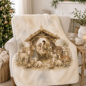 May include: A cream-coloured fleece throw blanket featuring a detailed watercolour-style illustration of a nativity scene. The print depicts a stable with figures, a cow, a donkey, several lambs, a deer, and a rabbit in soft brown and tan tones.