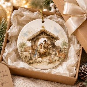 Puede incluir: Un adorno redondo de cerámica blanca con una detallada ilustración en acuarela de un belén con un establo, figuras y animales. El adorno cuelga de un cordón dorado y se presenta en una caja de regalo marrón con papel de seda blanco.