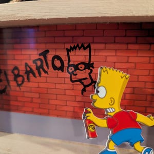 May include: A wooden framed art piece featuring Bart Simpson graffiti art. The artwork depicts Bart holding a spray can in front of a brick wall with the words "El Barto" and a stencil of Bart's face. The character is wearing a red shirt, blue shoes, and has yellow hair.
