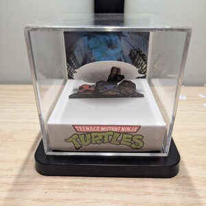 May include: A clear acrylic display case containing a Teenage Mutant Ninja Turtles diorama. The base features the green Teenage Mutant Ninja Turtles logo. Inside, a cutout shows three turtle characters emerging from a dark, city-themed background.