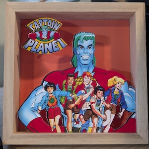 May include: A square wooden shadow box frame containing a layered paper cutout of Captain Planet and the Planeteers. The artwork features the blue-skinned hero behind five diverse characters, with the Captain Planet logo displayed in the upper left corner.