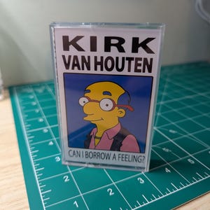 May include: A clear plastic cassette tape case featuring a printed insert with the text KIRK VAN HOUTEN and CAN I BORROW A FEELING? above and below a cartoon illustration of a yellow-skinned character with glasses, a pink shirt, and a black vest.