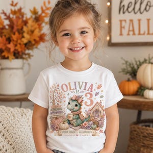 Puede incluir: Una camiseta blanca con un estampado gráfico colorido de un dinosaurio de dibujos animados con un lazo y una bufanda, sosteniendo una taza. El diseño incluye el texto OLIVIA IS 3, hojas de otoño, calabazas, una casa pequeña y LITTLE MOMENTS. BIG MEMORIES.
