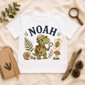 Puede incluir: Una camiseta blanca de manga corta con el gráfico de un dinosaurio explorador verde con sombrero de safari y mochila. El dinosaurio sostiene una lupa y un cuaderno. El nombre NOAH está impreso en azul oscuro sobre el dinosaurio, rodeado de ilustraciones de un helecho, un fósil, un hueso, una seta y una hoja sobre un papel.