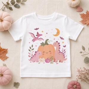 Puede incluir: Una camiseta blanca de manga corta con un estampado gráfico colorido de dos dinosaurios rosas, un pterodáctilo volador con sombrero, una calabaza naranja grande sonriente, calabazas más pequeñas, una luna creciente, murciélagos y estrellas con temática otoñal.