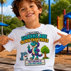 Puede incluir: Una camiseta blanca de manga corta con un estampado gráfico que incluye el texto "ACTUALLY... SHARKS ARE OLDER THAN TREES" y "FACT CHECKED BY A KID WHO KNOWS". El diseño muestra un tiburón, un dinosaurio morado y turquesa y un árbol.
