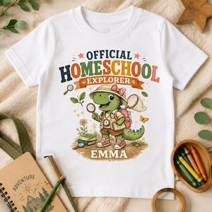 Puede incluir: Una camiseta blanca con un gráfico de un dinosaurio de dibujos animados vestido de explorador con lupa, mochila y red para mariposas. El texto dice OFFICIAL HOMESCHOOL EXPLORER y el nombre EMMA está impreso en letras grandes en la parte inferior.