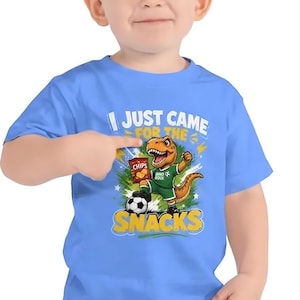 Puede incluir: Una camiseta azul claro con un dinosaurio de dibujos animados con uniforme de fútbol sosteniendo una bolsa de patatas fritas. La camiseta tiene el texto "I JUST CAME FOR THE SNACKS" en blanco y amarillo. El niño lleva vaqueros azules.