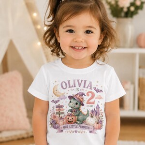 Puede incluir: Una camiseta blanca de manga corta con un estampado gráfico de un dragón verde con un sombrero de bruja morado, sosteniendo un cubo de calabaza. El texto dice OLIVIA IS 2 YEARS OUR LITTLE PUMPKIN, rodeado de murciélagos, una luna y un castillo.