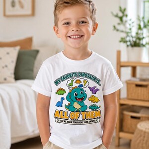 Puede incluir: Una camiseta blanca de manga corta con un gráfico colorido de un dinosaurio central de color verde azulado rodeado de dinosaurios más pequeños en burbujas de pensamiento. El texto dice: MY FAVORITE DINOSAUR IS... ALL OF THEM. ASK ME AGAIN TOMORROW, SAME ANSWER.
