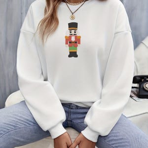 K&ouml;nnte beinhalten: Ein wei&szlig;es Langarm-Sweatshirt mit einer zentralen Grafik eines Nussknacker-Soldaten. Der Nussknacker ist mit einem schwarzen Hut, einer roten Jacke, einer gr&uuml;nen Hose und schwarzen Stiefeln dargestellt. Das Sweatshirt hat einen Rundhalsausschnitt.