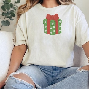 K&ouml;nnte beinhalten: Ein cremefarbenes Kurzarm-T-Shirt mit einer mittigen Grafik eines gr&uuml;nen Geschenkkartons mit wei&szlig;en Punkten und einer gro&szlig;en roten Schleife oben. Das Shirt hat einen Rundhalsausschnitt und eine lockere Passform mit festlichem Motiv.