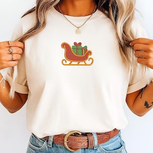 K&ouml;nnte beinhalten: Ein cremefarbenes Kurzarm-T-Shirt mit einer zentralen Stickerei eines roten Schlittens mit goldenen Kufen, der gr&uuml;ne Geschenkkartons mit roten Schleifen tr&auml;gt. Das T-Shirt wird mit einem braunen Lederg&uuml;rtel und einer Goldkette kombiniert.