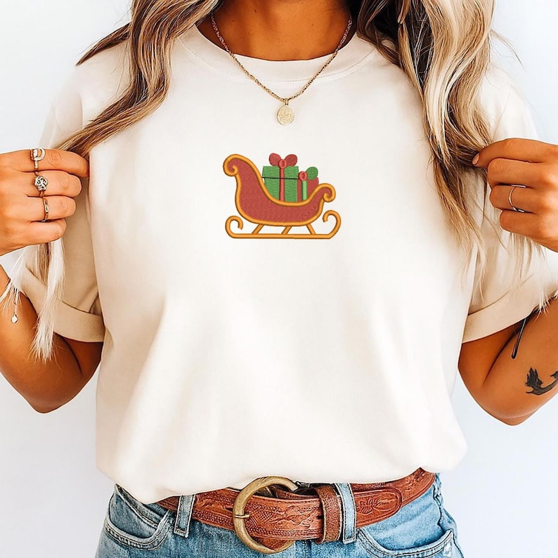 K&ouml;nnte beinhalten: Ein cremefarbenes Kurzarm-T-Shirt mit einer zentralen Stickerei eines roten Schlittens mit goldenen Kufen, der gr&uuml;ne Geschenkkartons mit roten Schleifen tr&auml;gt. Das T-Shirt wird mit einem braunen Lederg&uuml;rtel und einer Goldkette kombiniert.