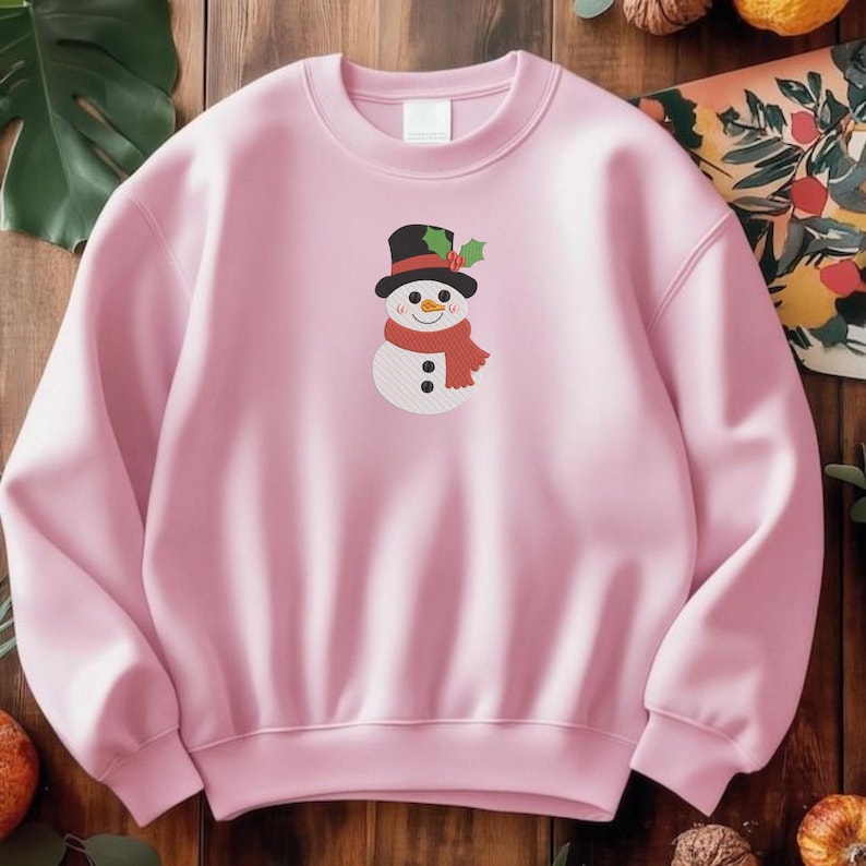 K&ouml;nnte beinhalten: Ein hellrosa Sweatshirt mit Rundhalsausschnitt und einem mittigen Motiv eines Schneemanns, der einen schwarzen Zylinder mit gr&uuml;nen Stechpalmenbl&auml;ttern und roten Beeren, einen roten Schal, eine orangefarbene Karottennase und zwei Kn&ouml;pfe tr&auml;gt.