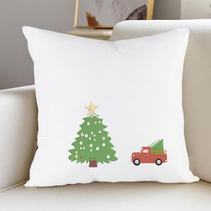 K&ouml;nnte beinhalten: Ein wei&szlig;es quadratisches Dekokissen mit einem gr&uuml;nen Weihnachtsbaum, der mit wei&szlig;en Ornamenten und einem goldenen Stern an der Spitze verziert ist, neben einem roten Pickup, der einen kleinen gr&uuml;nen Tannenbaum auf der Ladefl&auml;che tr&auml;gt.