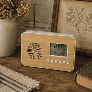 May include: A retro-style digital clock radio with a light wood grain finish and white trim. The clock displays the time, date, and temperature. It has a speaker, buttons, and a retractable antenna. The clock is on a wooden surface.