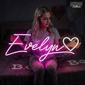 Puede incluir: Un letrero de neón rosa brillante con el nombre "Evelyn" escrito en cursiva y un corazón. La persona sostiene el letrero. El letrero está sobre una cama con la palabra "Be" visible.