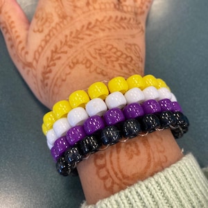 May include: A stack of beaded bracelets in the non-binary pride flag colors: yellow, white, purple, and black. The bracelets are worn on a wrist with henna designs. The bracelets are made of plastic beads strung together.