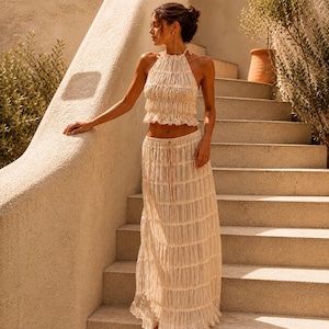 Könnte beinhalten: Cremefarbenes zweiteiliges Outfit mit Neckholder-Top und bodenlangem Rock. Das Oberteil hat einen gerüschten Ausschnitt und einen verkürzten Schnitt. Der Rock hat ein vertikales Falten-Design. Das Outfit besteht aus leichtem, fließendem Stoff.