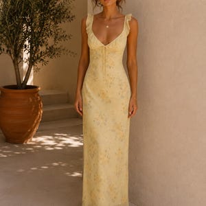 May include: A long, yellow floral print dress with a V-neckline and ruffled straps. The dress has a flowing silhouette and a subtle floral pattern in shades of yellow and blue. The dress is made of a lightweight fabric.