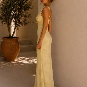 May include: A yellow floral print maxi dress with spaghetti straps and a ruffled neckline. The dress has a fitted bodice and a flowing skirt. The model is standing next to a potted olive tree. The dress is made of a lightweight fabric.