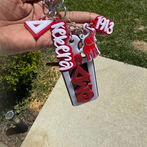 May include: A collection of red, white, and black keychains. The keychains include a triangle, the word "Kekera", the number "1913", an elephant, and Greek letters. The keychains are attached to a silver keyring.