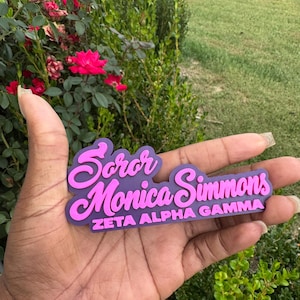 May include: A custom name badge featuring the text Soror Monica Simmons in a pink cursive font layered over a purple background, with the text ZETA ALPHA GAMMA printed in a bold pink sans-serif font on the bottom line of the badge.