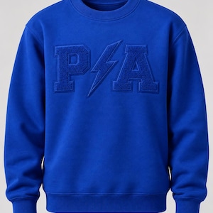 May include: A vibrant royal blue crewneck sweatshirt featuring a textured, embossed graphic on the chest. The design displays the letters P and A in a collegiate font, separated by a central lightning bolt symbol, all in a matching blue tone.