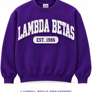 Peut inclure: Un sweat-shirt à col rond violet profond avec le texte brodé blanc LAMBDA BETAS dans une police universitaire arquée, un petit écusson ovale blanc en dessous affichant EST. 1986 en violet, et un petit logo ΣΛΒ sur l'étiquette du col.