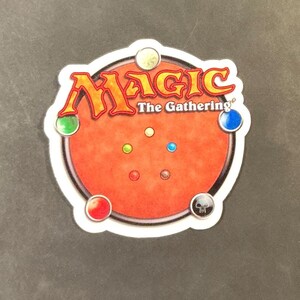 May include: A circular Magic: The Gathering playmat with a red center and the game's logo in orange and yellow. The mat features colored circles around the edge, representing different mana types, and a skull icon.