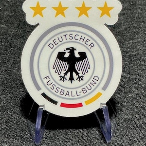 May include: A circular sticker featuring the Deutscher Fussball-Bund logo with a black eagle in the center, surrounded by text, four yellow stars at the top, and a black, red, and yellow horizontal stripe at the bottom, displayed on a stand.