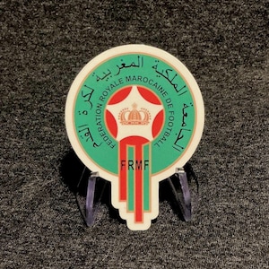 May include: A circular sticker featuring the Royal Moroccan Football Federation logo. It displays a central red and white emblem with a crown, surrounded by a green border with Arabic text and the English text Federation Royale Marocaine de Football.