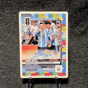 May include: A Panini Instant trading card featuring Lionel Messi in an Argentina jersey, with a blue ink autograph across the center. The card displays the text FIFA World Cup 2026, Lionel Messi, Argentina legend sets FIFA World Cup goals mark, and June 22, 2026.
