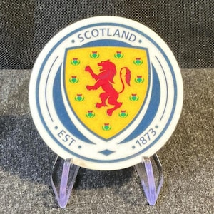 May include: A circular ceramic coaster featuring the Scotland crest with a red lion on a yellow shield, surrounded by small green thistle icons. The border is blue and white with the text SCOTLAND and EST 1873 printed in a bold, dark blue font.