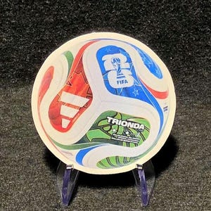 May include: A round, flat, circular coaster featuring a graphic of a soccer ball with red, blue, and green panels. The design includes the Adidas logo, the FIFA World Cup trophy icon, and the text TRIONDA, along with smaller star patterns.