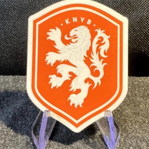 May include: A shield-shaped sticker featuring an orange background with a white lion graphic and the text KNVB at the top. The sticker is displayed on a clear plastic stand, showcasing the emblem of the Royal Dutch Football Association.