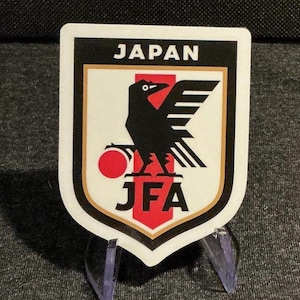 May include: A shield-shaped sticker featuring the Japan Football Association emblem. It displays a black three-legged crow over a red vertical stripe, a red circle, and the text JAPAN and JFA in black lettering against a white and gold background.