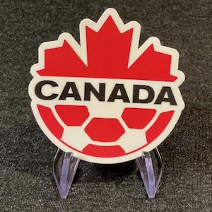 May include: A die-cut sticker featuring a red maple leaf at the top and a red and white soccer ball pattern at the bottom. The word CANADA is printed in bold black capital letters across the center of the sticker on a white background.