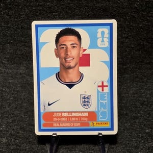 May include: A rectangular Panini trading card featuring a portrait of Jude Bellingham in a white England soccer jersey. The card includes the text Jude Bellingham, 29-6-2003, 1.86 m, 75 kg, Real Madrid CF (ESP), and a FIFA logo on a blue background.