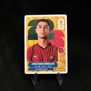 May include: A rectangular Panini trading card featuring a portrait of Cristiano Ronaldo wearing a red jersey. The card displays the text CRISTIANO RONALDO, 5-2-1985, 1.87 m, 83 kg, and AL-NASSR FC (KSA) against a yellow background with a large green and red 23.