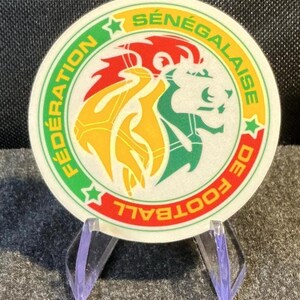 May include: A circular sticker featuring the logo of the Fédération Sénégalaise de Football. The design displays a stylized lion head in yellow, green, and red, surrounded by text and two green stars on a white background with a thin red border.