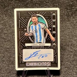 May include: A rectangular sports trading card featuring a portrait of Lionel Messi in an Argentina soccer jersey. The card has a black background with geometric lines, the text AURORA, LIONEL MESSI, ARGENTINA, and OBSIDIAN, and a blue ink autograph.